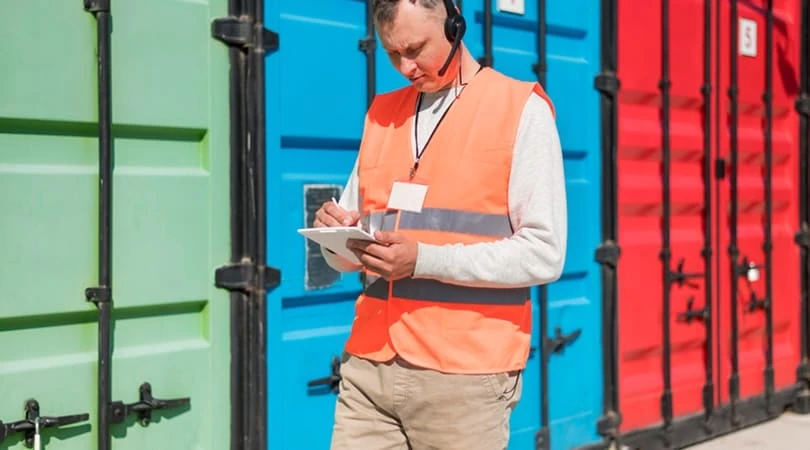 Un homme en gilet orange et écouteurs se tient devant des conteneurs de transport.